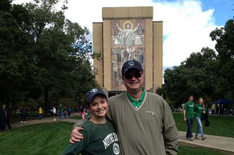 A man and a young girl wearing Notre Dame apparel stand together on a campus lawn, smiling with their arms around each other, with a large building featuring a religious mural visible in the background.