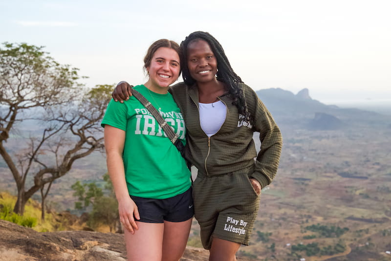 Two women standing arm in arm on a scenic overlook, smiling at the camera, with a wide valley and distant mountains in the background. One wears a green T-shirt and black shorts, and the other wears a dark green hoodie and matching shorts.