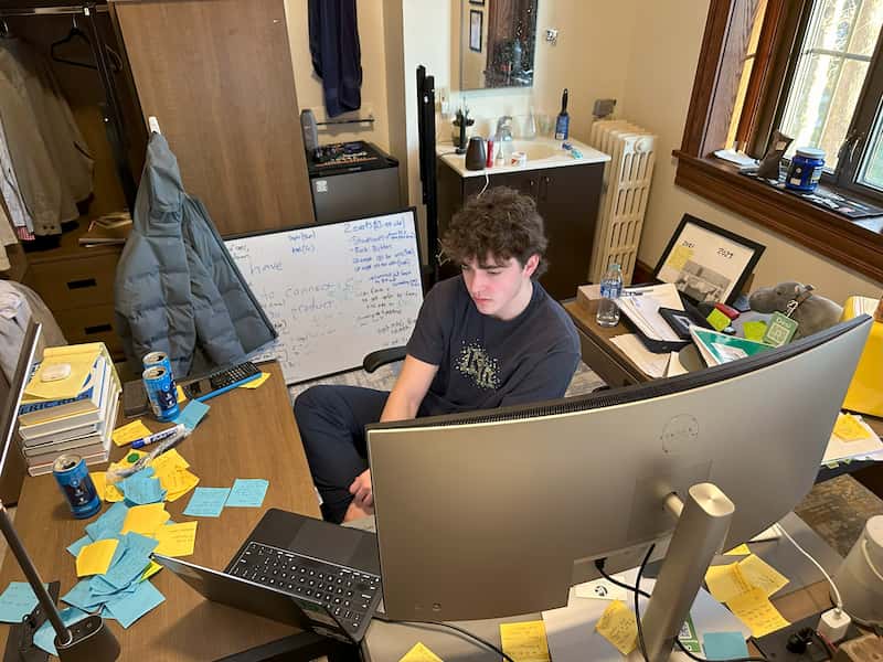 A young person sits at a wooden desk in a small, dorm-like room, focused on a large curved computer monitor. The desk is cluttered with blue and yellow sticky notes, notebooks, pens, and several empty energy drink cans. A laptop sits open beside the monitor. Behind them, a whiteboard covered in handwritten notes leans against a cabinet next to a mini fridge and a small sink. A window on the right lets in natural light, and the compact space appears busy and well-used for studying or work.