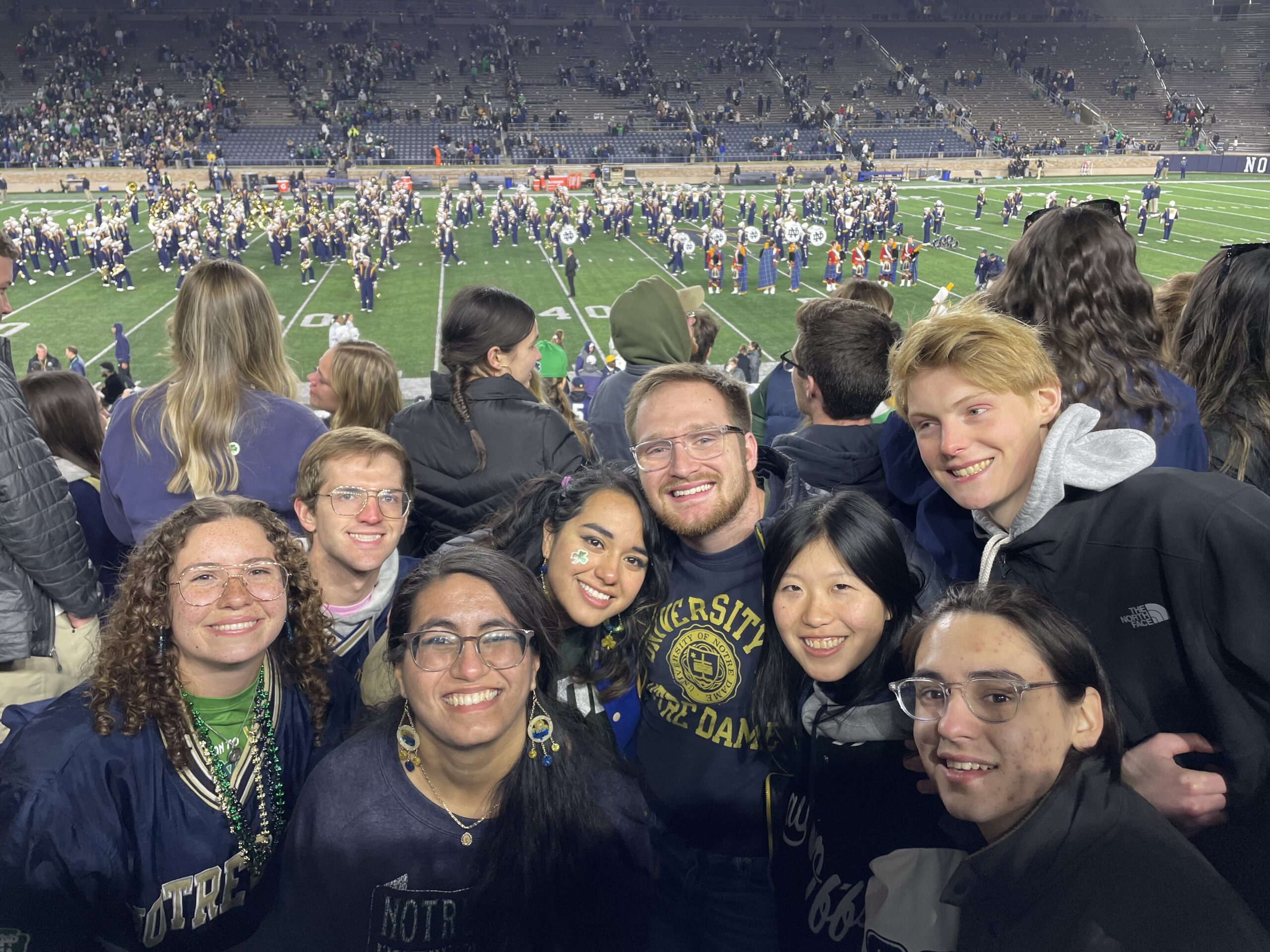 A group of friends smiling for a photo in the stands of a football stadium at night, with a marching band performing on the field below and stadium seating filled with spectators in the background.