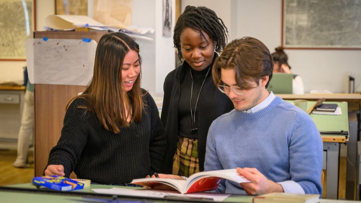 Three students in a studio facility standing behind a table looking at the same book