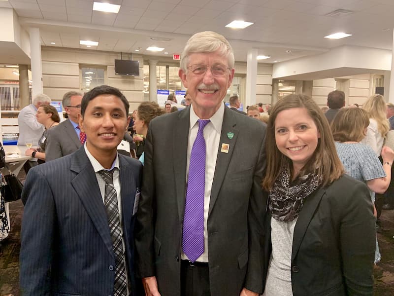 Three people dressed in business attire stand together smiling at a crowded indoor networking event, with other attendees mingling in the background.