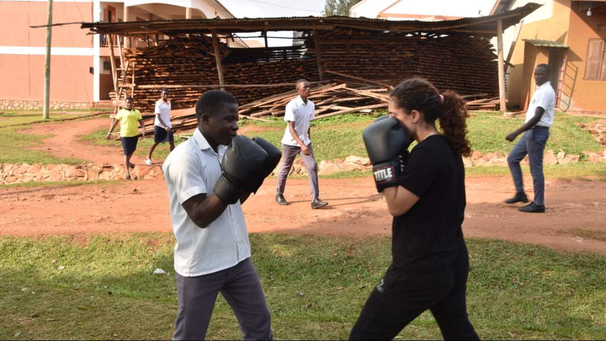 Two people standing outside, facing one another while holding their hands near their faces, wearing black boxing gloves