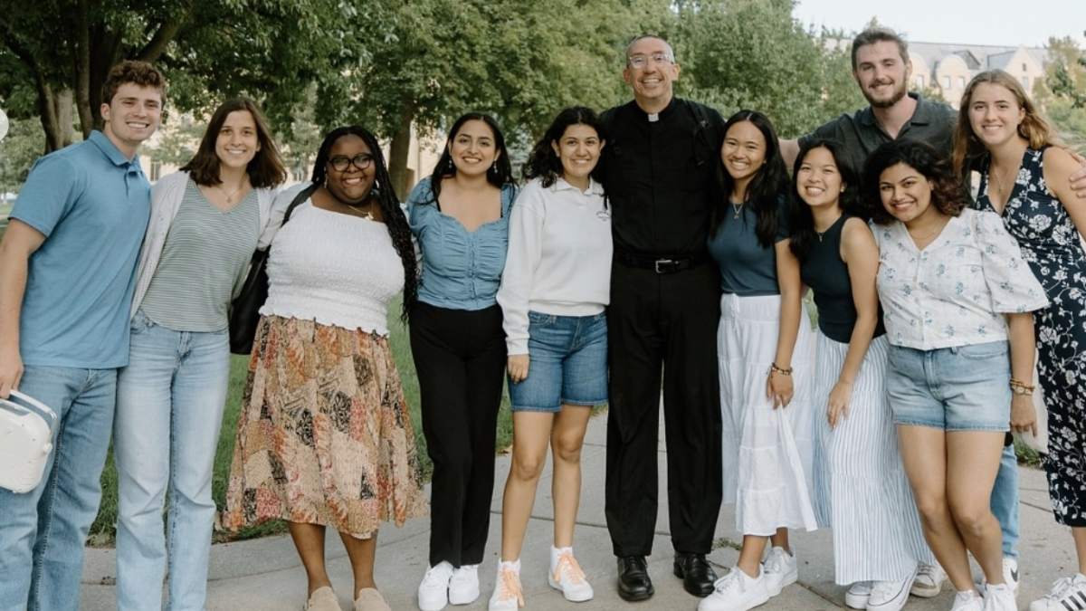 Group of 10 students standing outside on a sidewalk in a group with Rev. Pete McCormick, C.S.C.