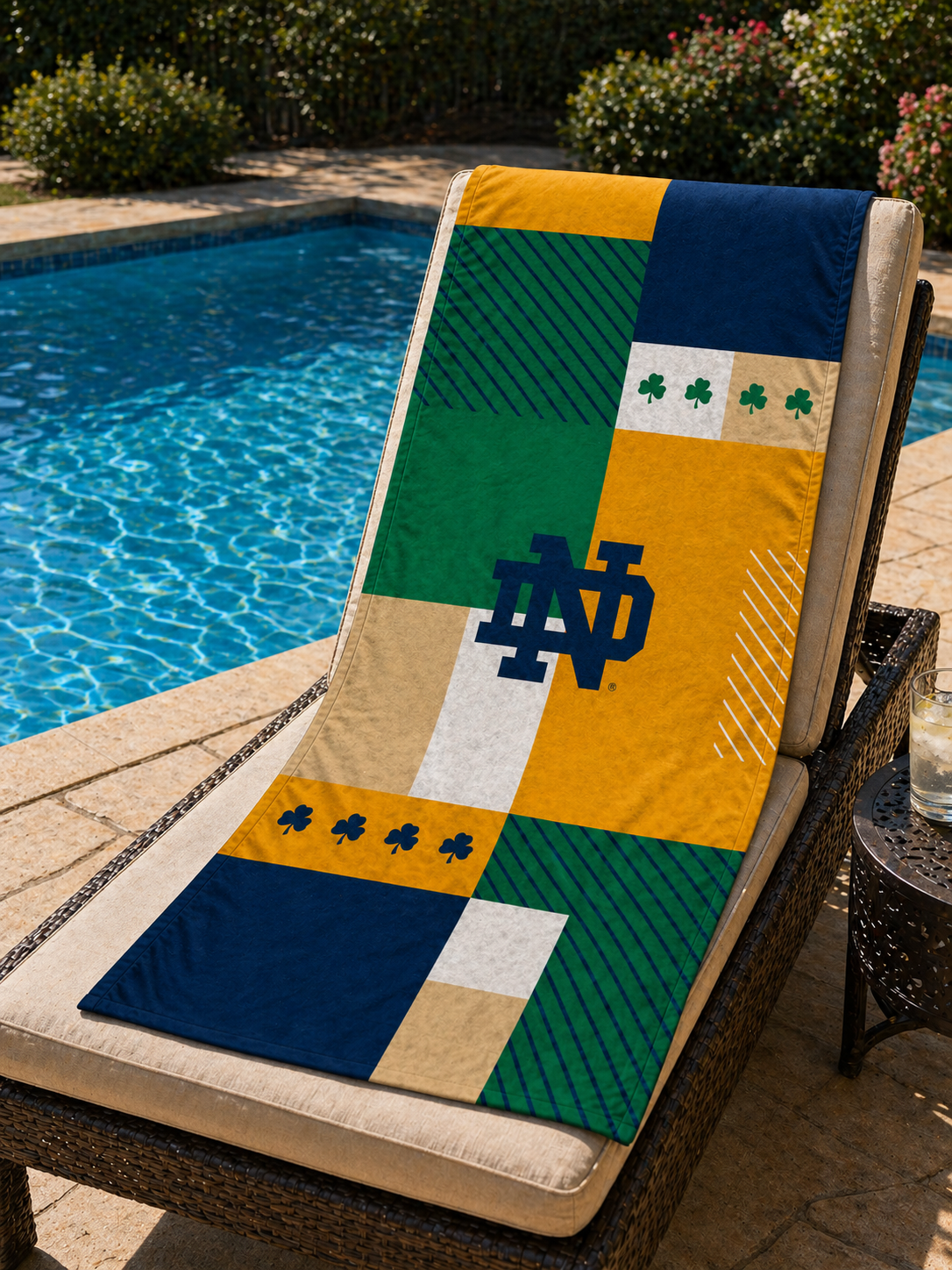 The 2026 Notre Dame beach towel, featuring a collage of gold, blue, green, and white squares adorned with a blue ND Monogram and multiple shamrocks, is draped over the back of a lounge chair in front of a swimming pool.