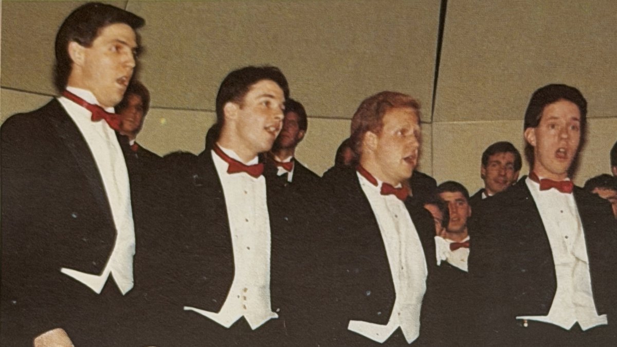 Four men standing in tuxedos with red bow ties, looking to the right while singing