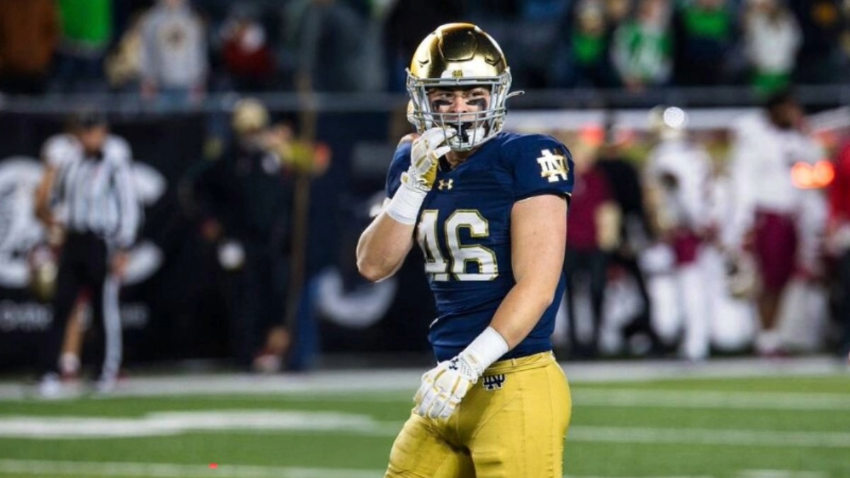 Jerry Rullo standing on a football field wearing gold pants, a dark blue Notre Dame jersey, and gold helmet, with a blurry crowd and referee in the back