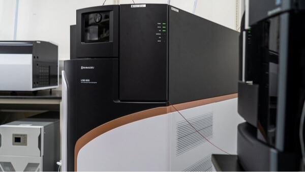 Close-up of a laboratory mass spectrometer, which looks like a black, brown, and gray rectangle, with connected tubing and adjacent black analytical equipment.