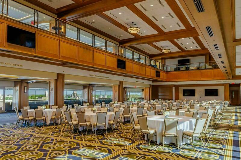 Large banquet hall with high wood-paneled ceilings, patterned carpet, and numerous round tables draped in white linens and surrounded by upholstered chairs. Natural light enters through large windows along one side of the room, and a second-level balcony overlooks the event space, creating an elegant setting for conferences, receptions, or formal gatherings.