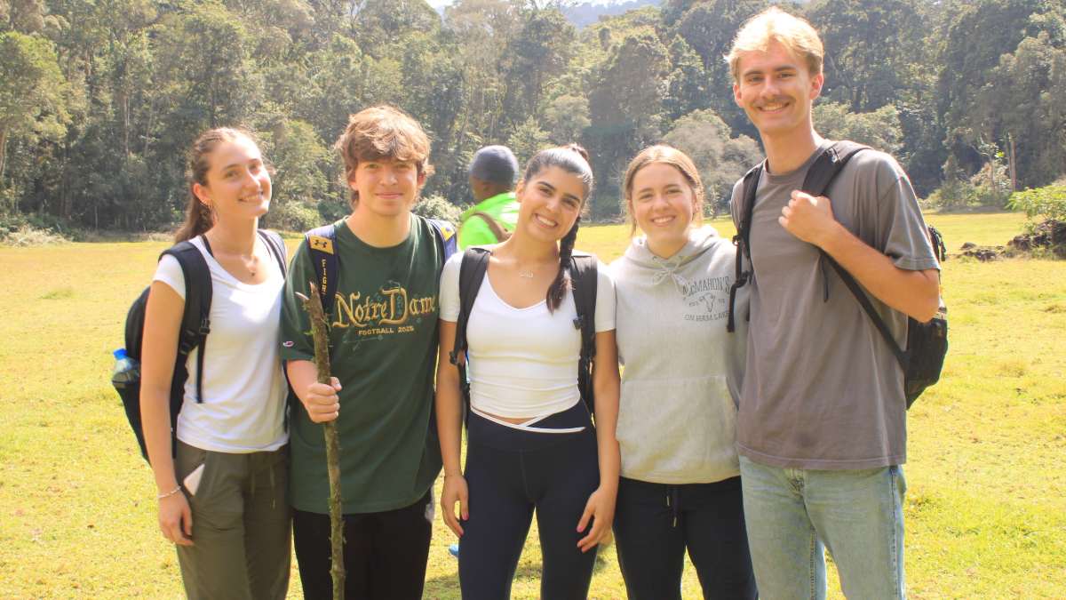 Five students standing together smiling, four wearing t-shirts and one in a sweatshirt, all wearing backpacks, outside in a field with trees in the background