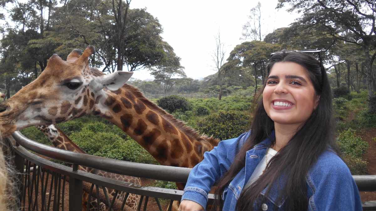 Woman wearing a jean jacket standing in the foreground smiling with sunglasses on her head, leaning on a railing with two giraffes behind her, one close to her arm