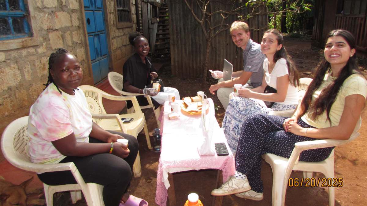 Five people sitting around a table with a pink tablecloth, holding sandwiches and cups, looking at the camera