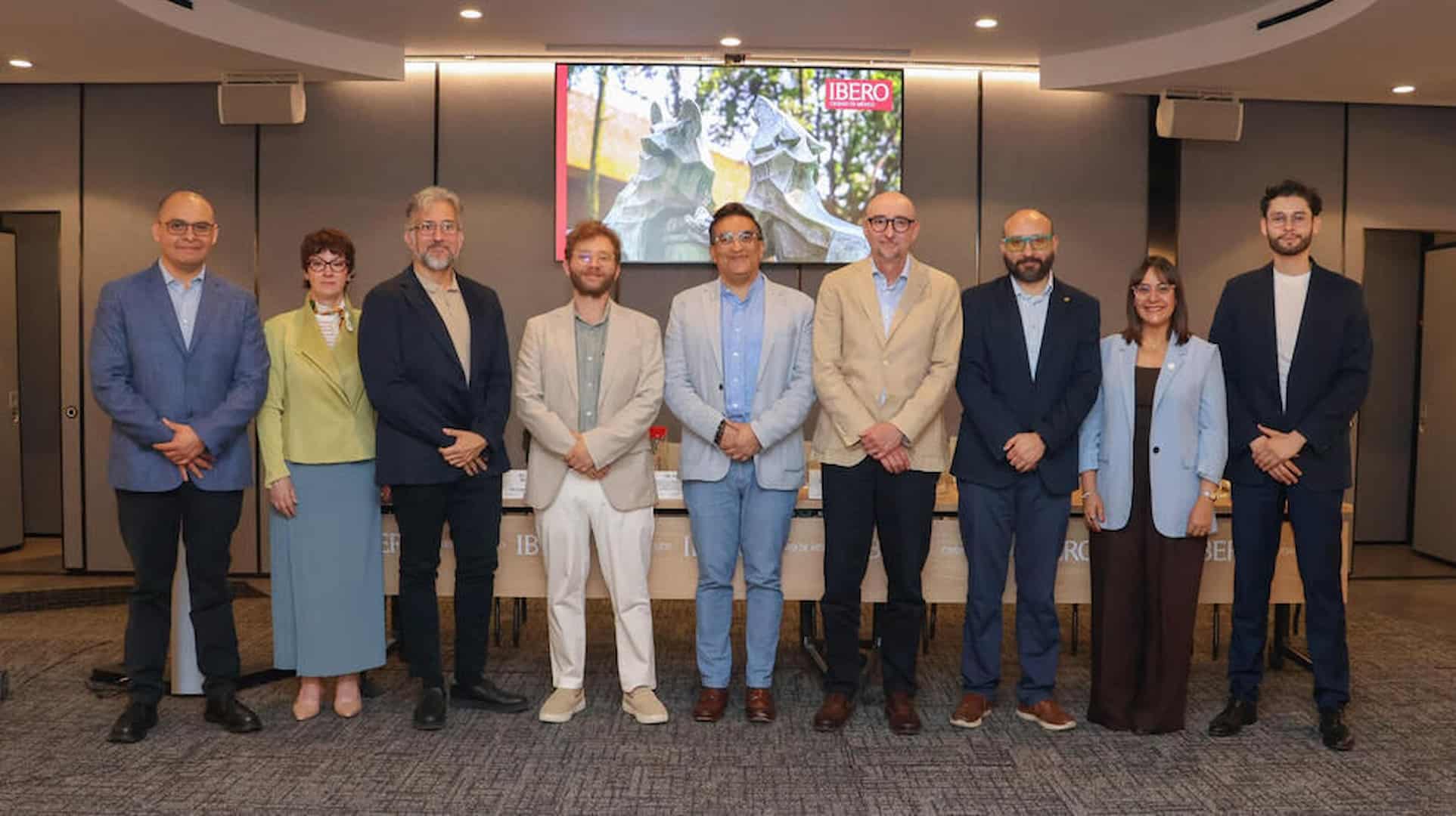 A group of 9 well-dressed academics pose together in front of a wide conference panel table, podium, and backdrop labeled IBERO.