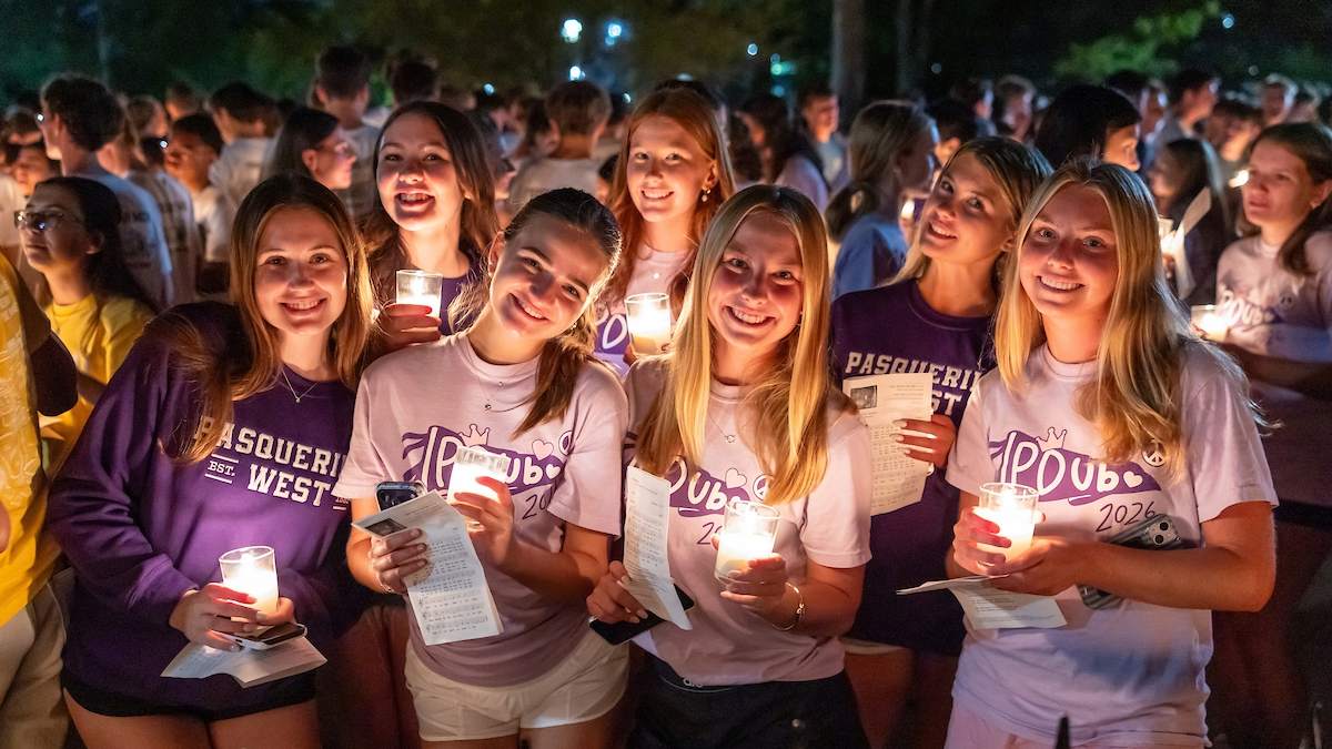 Group of seven female Notre Dame students standing together, holding candles, wearing purple and white residence hall t-shirts that say Pasquerilla West and PDub 2026.