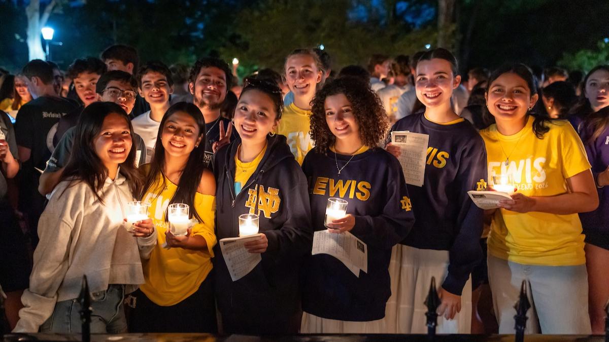 A group of Notre Dame students gathered outside in candlelight, smiling at the camera, wearing Lewis Hall and Notre Dame gear