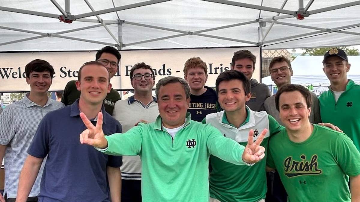 Group of 11 men, standing together under a tent, wearing Notre Dame gear, Fr. Ralph in front holding up two peace signs